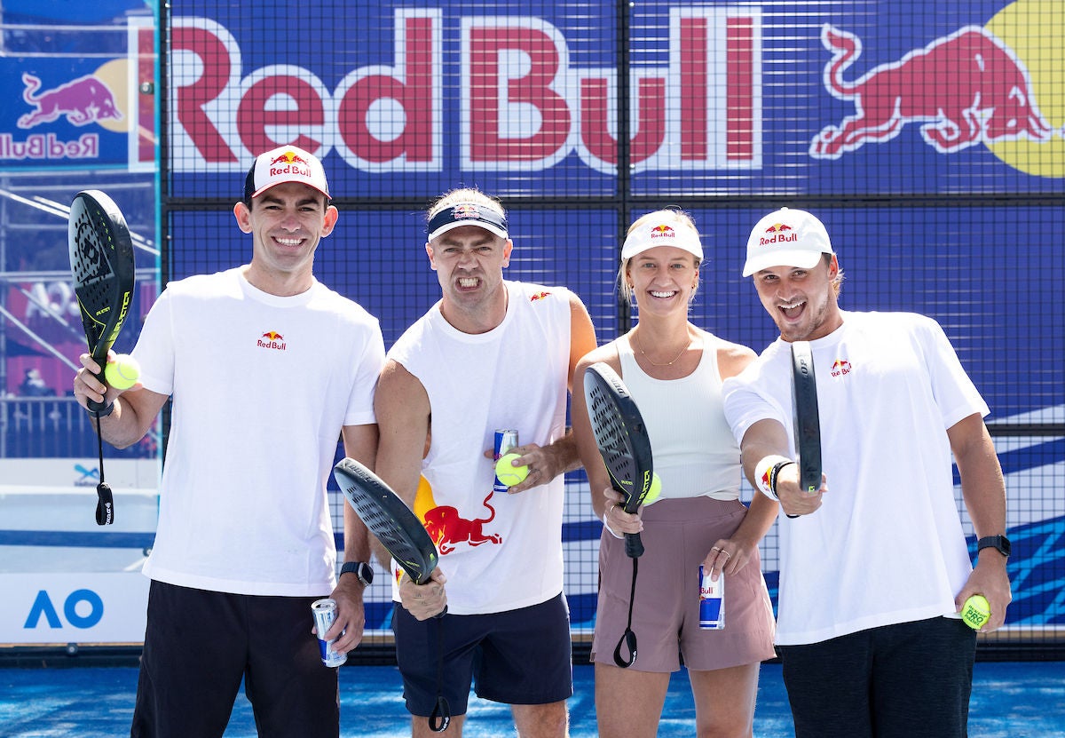 Four padel players wearing white t-shirts on a padel court with a blue hoarding behind them.