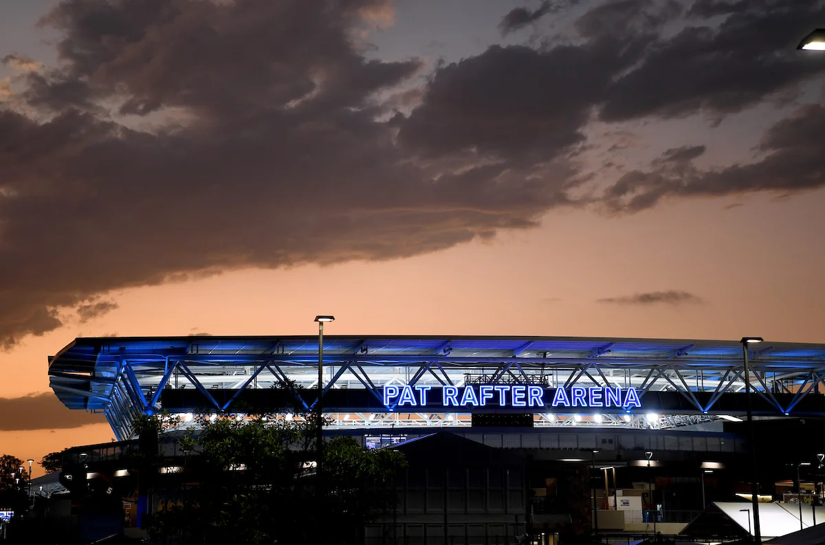 A stadium lit up with flood lights in front of a mood sky. A blue sign saying Pat Rafter Arena is lit up.