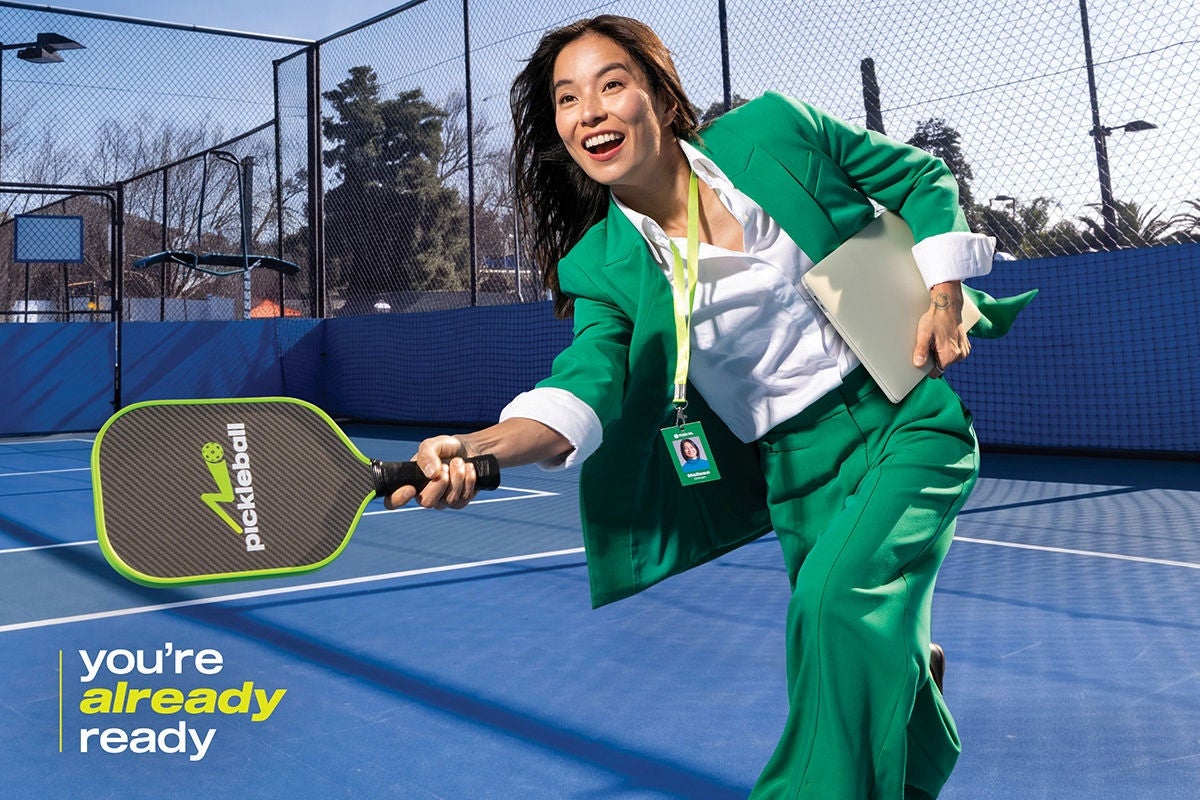 A smiling woman in a bright green suit lunges forward on a blue pickleball court, holding a paddle and a laptop under her arm.