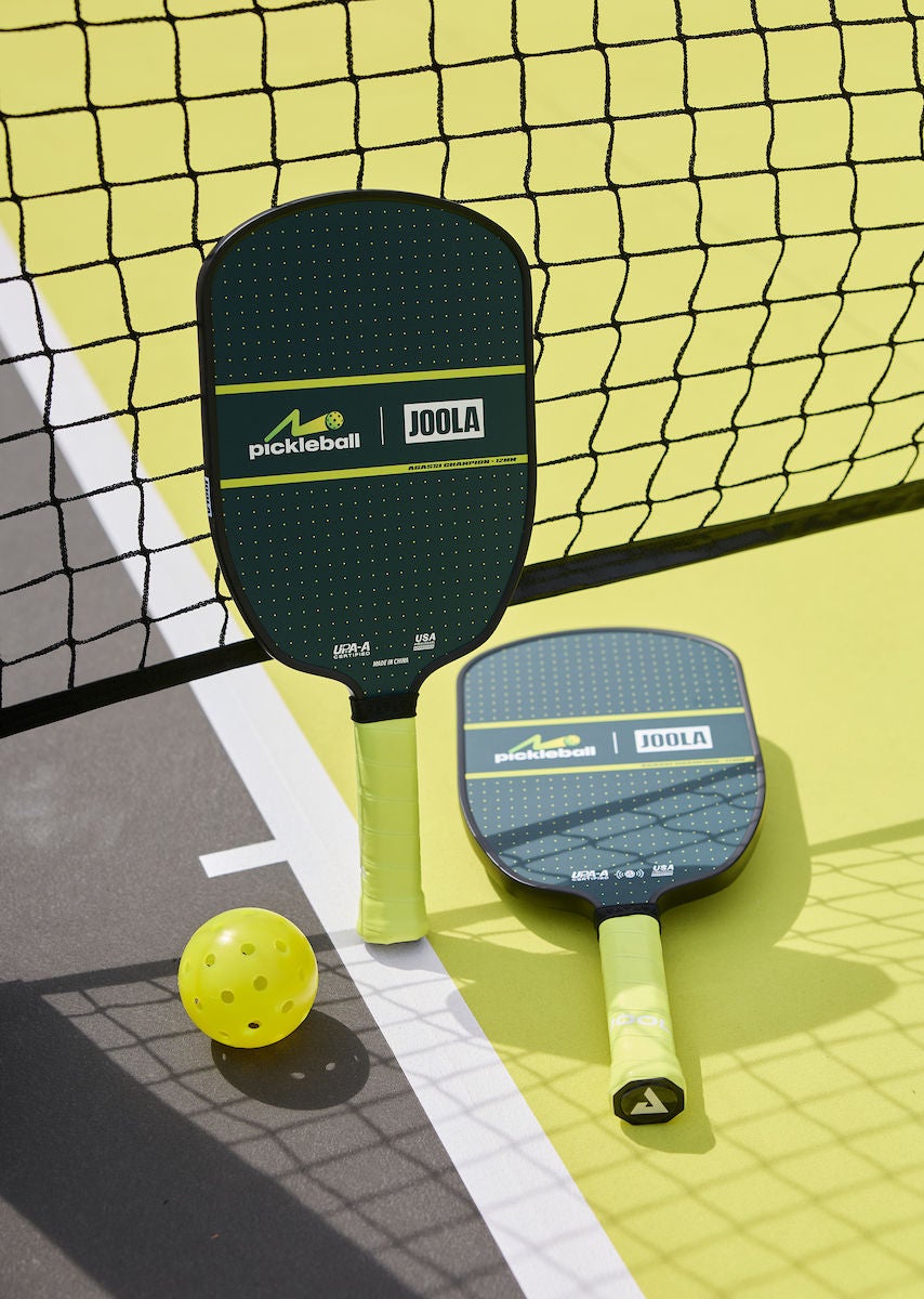 A pickleball padel and ball leaning against a net on a yellow court surface.